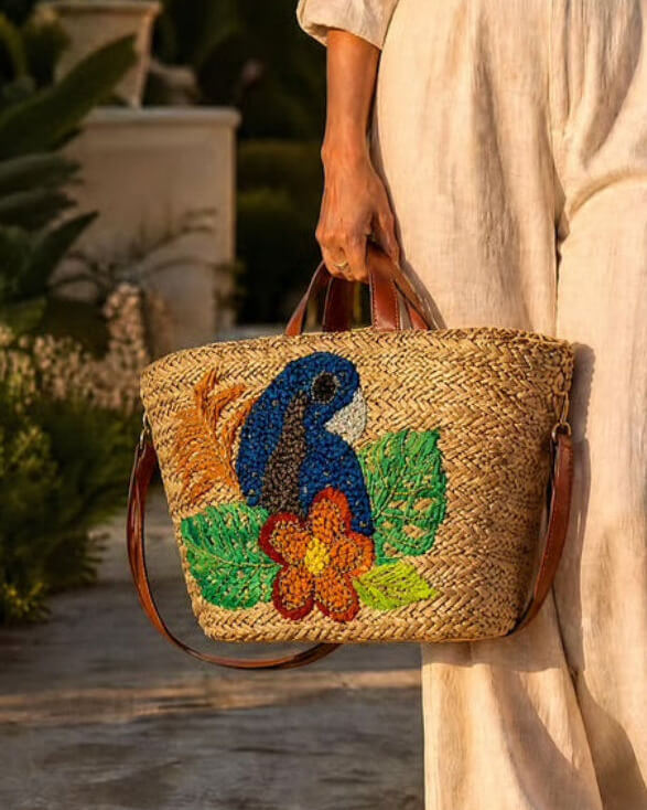 A woven straw tote bag with a colorful parrot and floral design, held by a person wearing a cream linen dress in an outdoor garden setting.