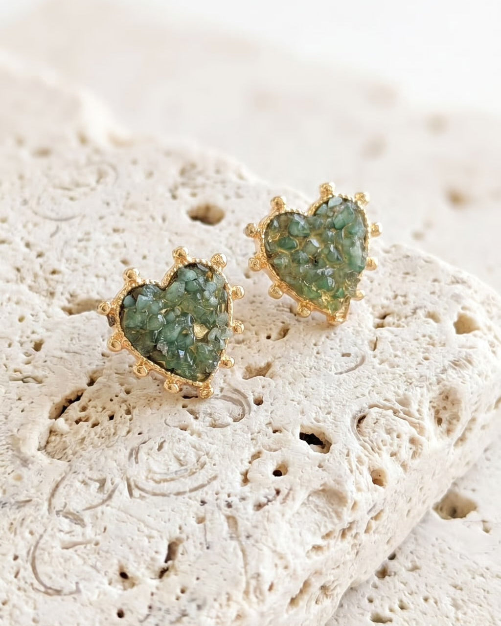 A pair of gold heart-shaped stud earrings with green glittery resin centers, resting on a textured beige stone tile against a soft white background.