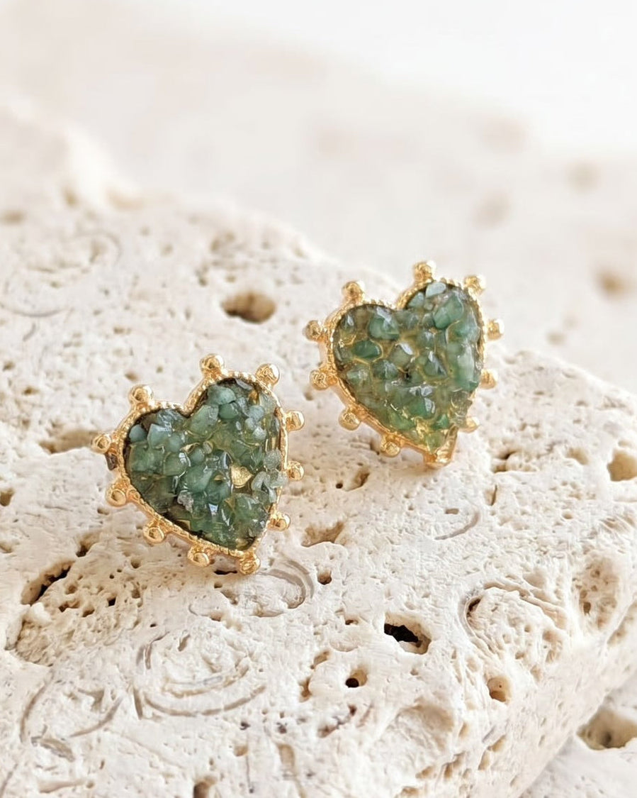 A pair of gold heart-shaped stud earrings with green glittery resin centers, resting on a textured beige stone tile against a soft white background.