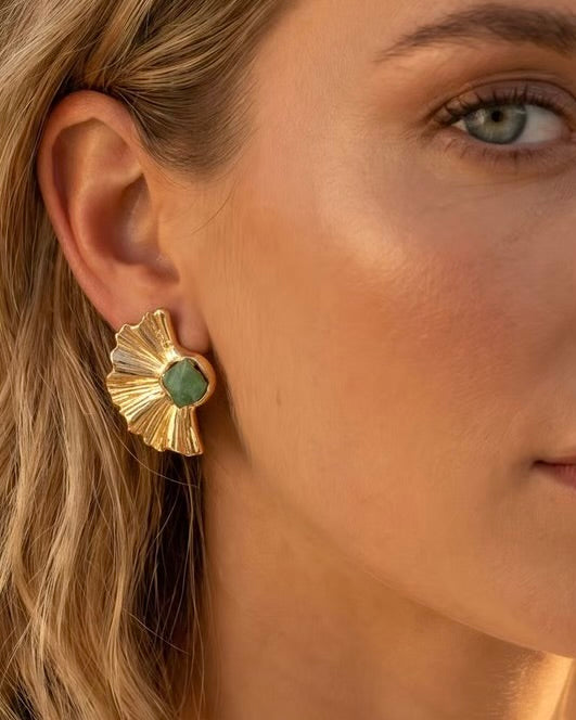 A close-up of a woman's ear wearing a gold fan-shaped stud earring with a green oval center stone.