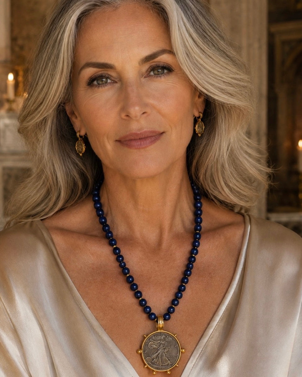 A close-up portrait of a woman with gray hair wearing a dark blue beaded necklace with a large gold-toned coin pendant, along with gold earrings and a cream satin top.
