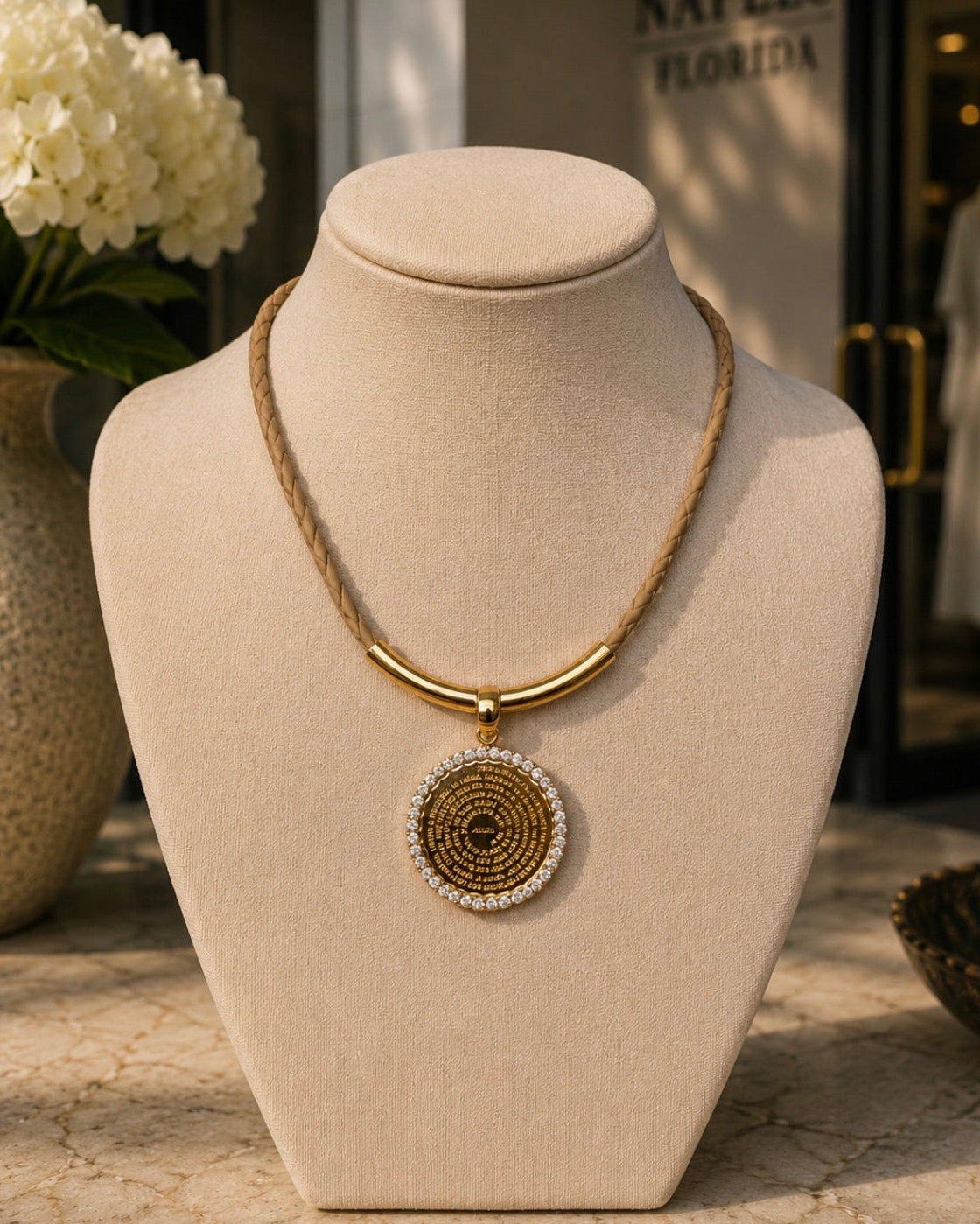 A gold and brown beaded necklace with a round pendant displayed on a beige jewelry bust. The necklace features a braided brown cord, a polished gold bar, and a circular pendant with brown woven details and a border of small clear stones.
