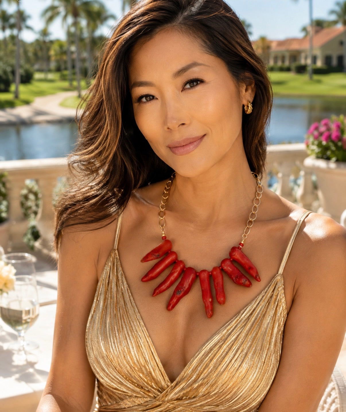 A woman wearing a gold metallic spaghetti-strap dress and a chunky red chili pepper statement necklace, posing outdoors with a pool and palm trees in the background.