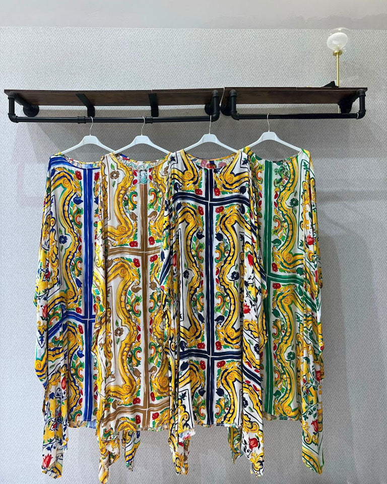 Three long-sleeve kaftan-style dresses with vibrant, colorful Mediterranean-inspired baroque and floral patterns in yellow, green, blue, and red, hanging on white hangers from a dark wooden wall rack.