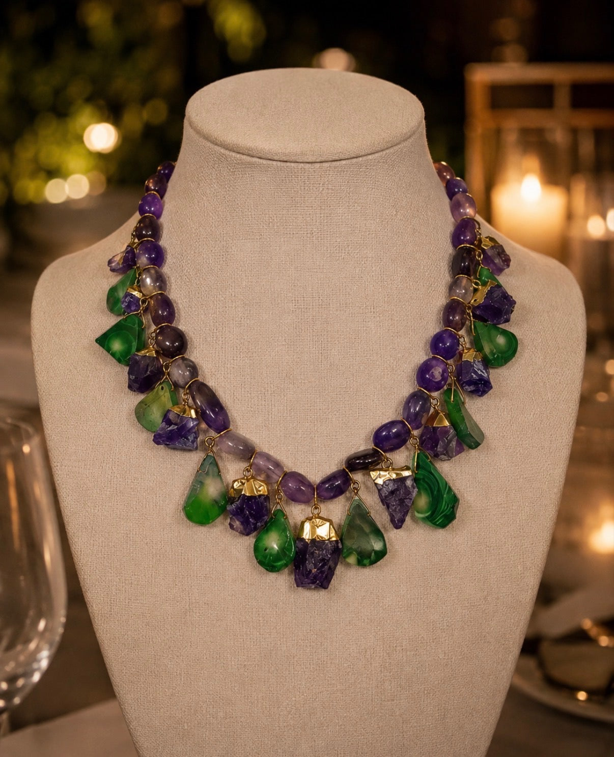 A statement necklace made of purple and green gemstone chips with gold-toned metal accents, displayed on a beige fabric jewelry bust against a softly blurred warm background.