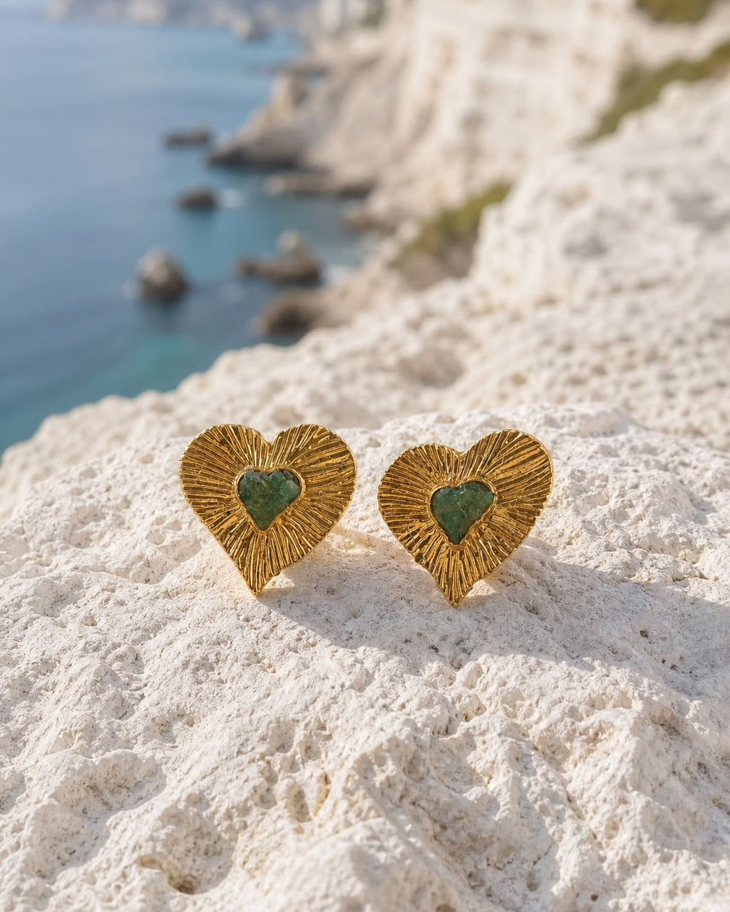 A pair of gold heart-shaped stud earrings with green inlaid centers, resting on a white rocky cliffside with a blue sea and distant land in the background.