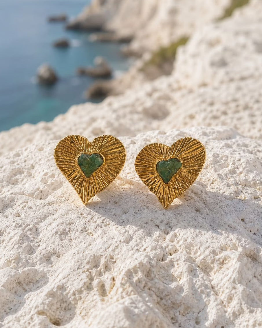A pair of gold heart-shaped stud earrings with green inlaid centers, resting on a white rocky cliffside with a blue sea and distant land in the background.