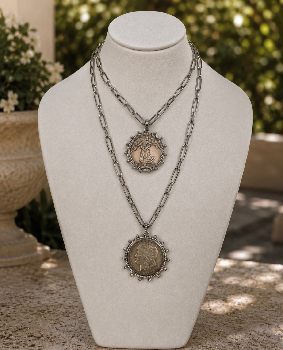 A double-strand silver-tone necklace displayed on a cream jewelry bust. The necklace features two circular coin-style pendants with ornate silver borders; the top pendant is smaller and the bottom pendant is larger, both showing detailed antique medallion designs.