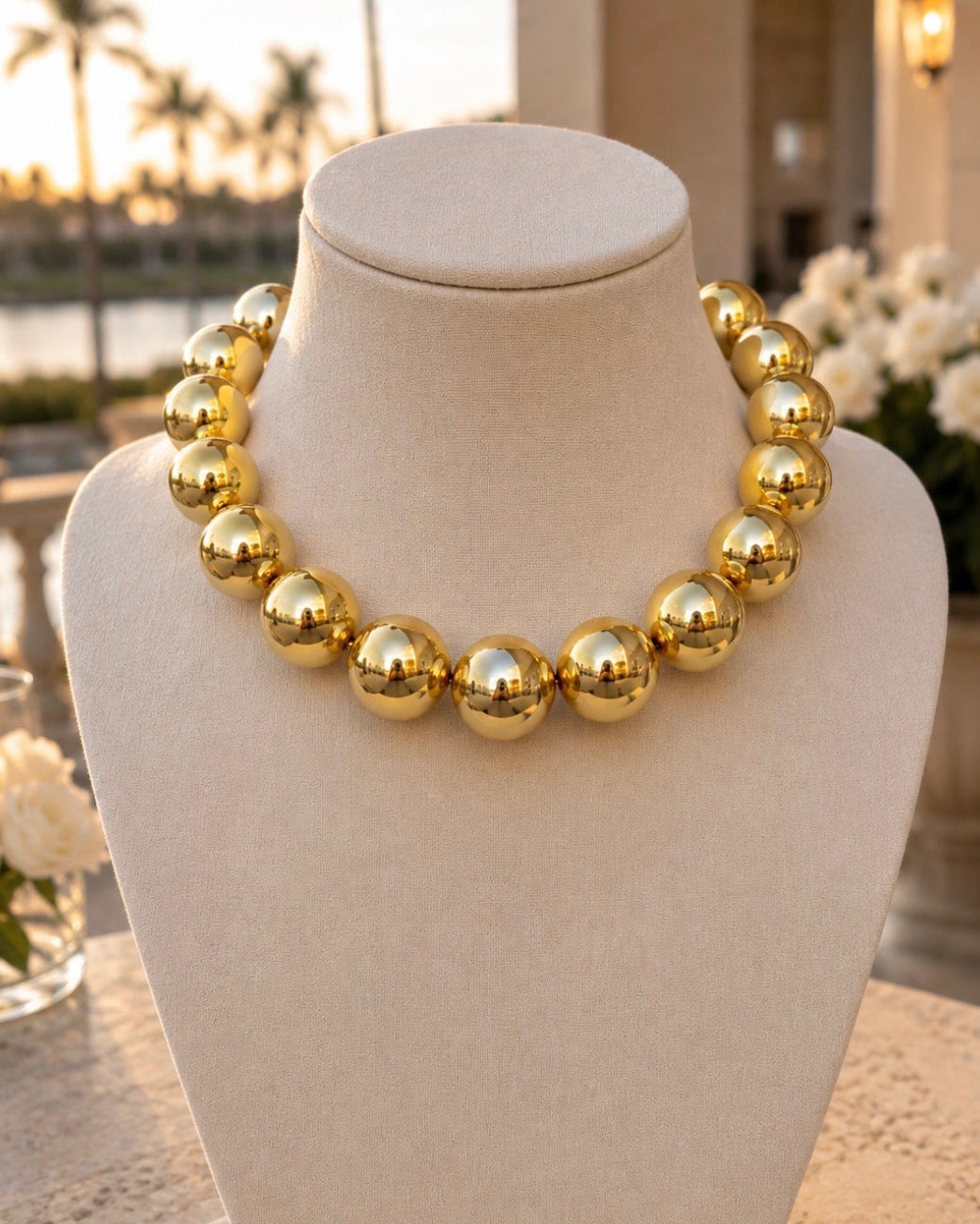 A chunky gold beaded necklace displayed on a cream-colored jewelry bust, set against a softly blurred outdoor patio background with flowers and palm trees.