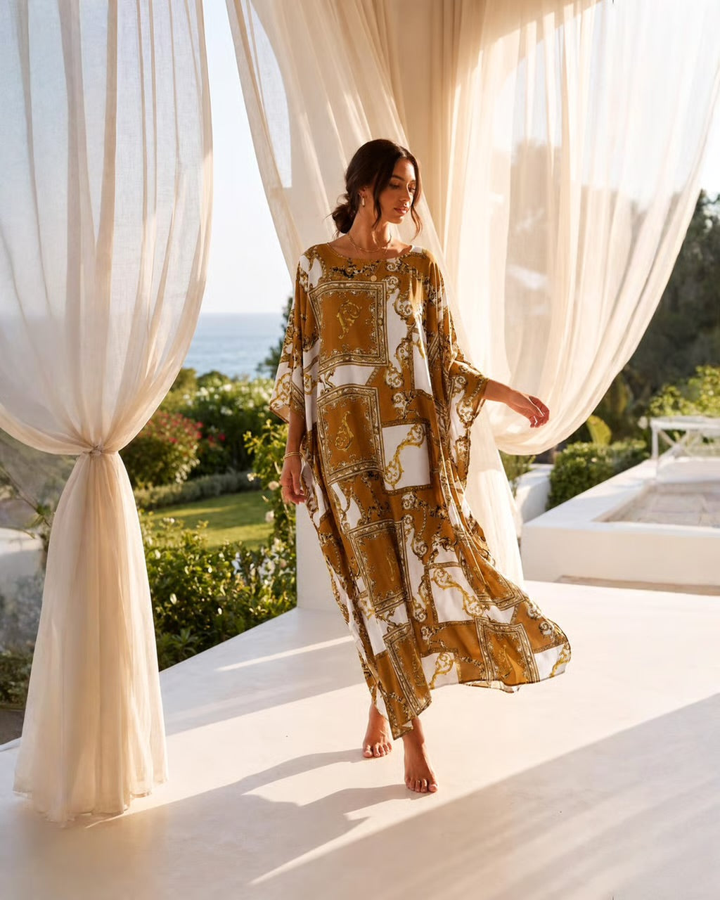 A woman wearing a flowing kaftan dress walks barefoot on a sunlit terrace framed by sheer cream drapes, with the ocean and greenery visible in the background. The dress has a warm brown and white ornate pattern.