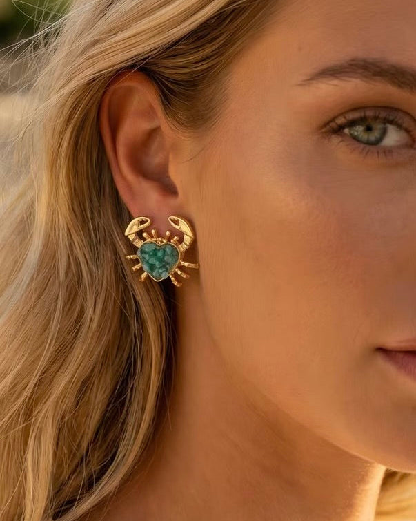 A close-up of a woman wearing a gold crab-shaped stud earring with a turquoise heart-shaped center, set against a softly blurred outdoor background.