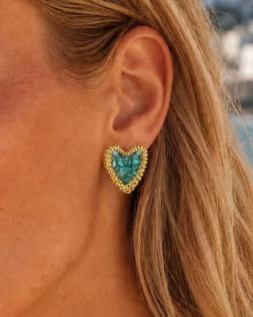A close-up of a woman's ear wearing a gold heart-shaped stud earring with a turquoise blue center stone, set against a blurred outdoor background.