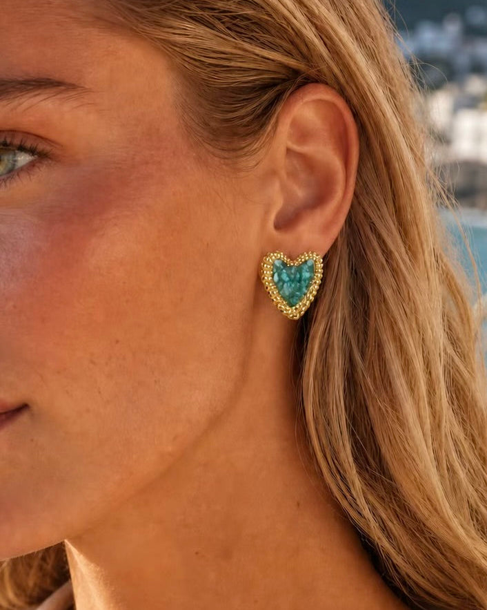 A close-up of a woman's ear wearing a gold heart-shaped stud earring with a turquoise blue center stone, set against a blurred outdoor background.