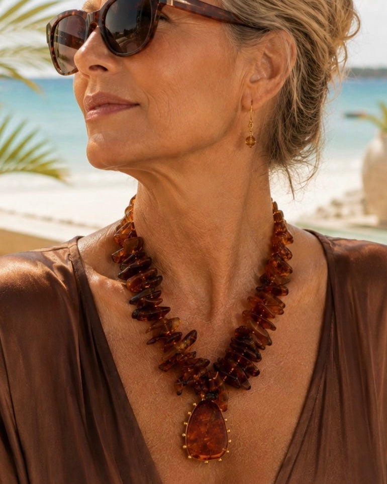 A woman wearing a chunky amber-brown stone necklace with a teardrop pendant, styled with sunglasses and a brown top, set against a sunny beach background.