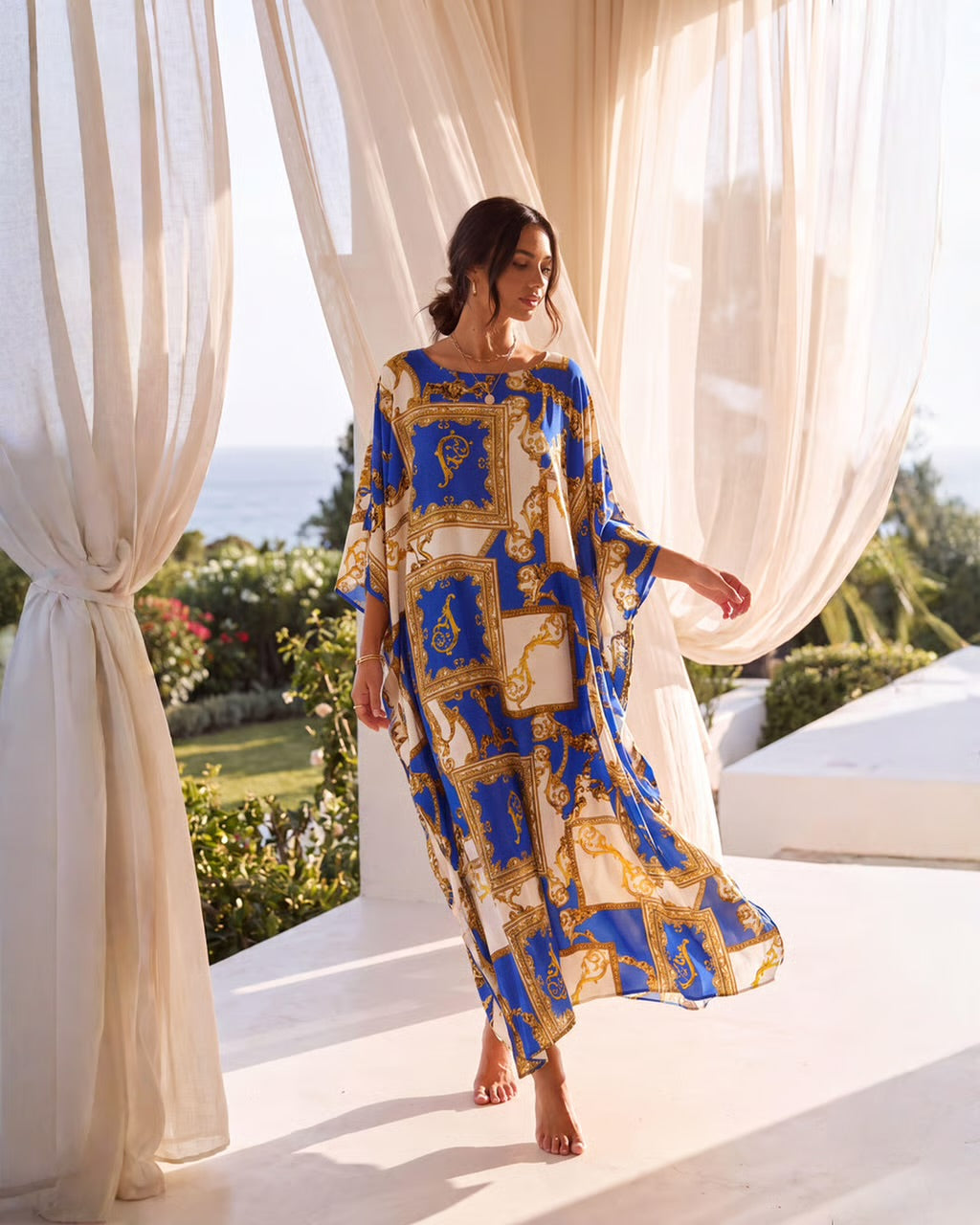 A woman wearing a flowing kaftan dress with a blue, gold, and white baroque-style pattern, standing barefoot on a white terrace framed by sheer cream curtains with a garden and ocean view in the background.
