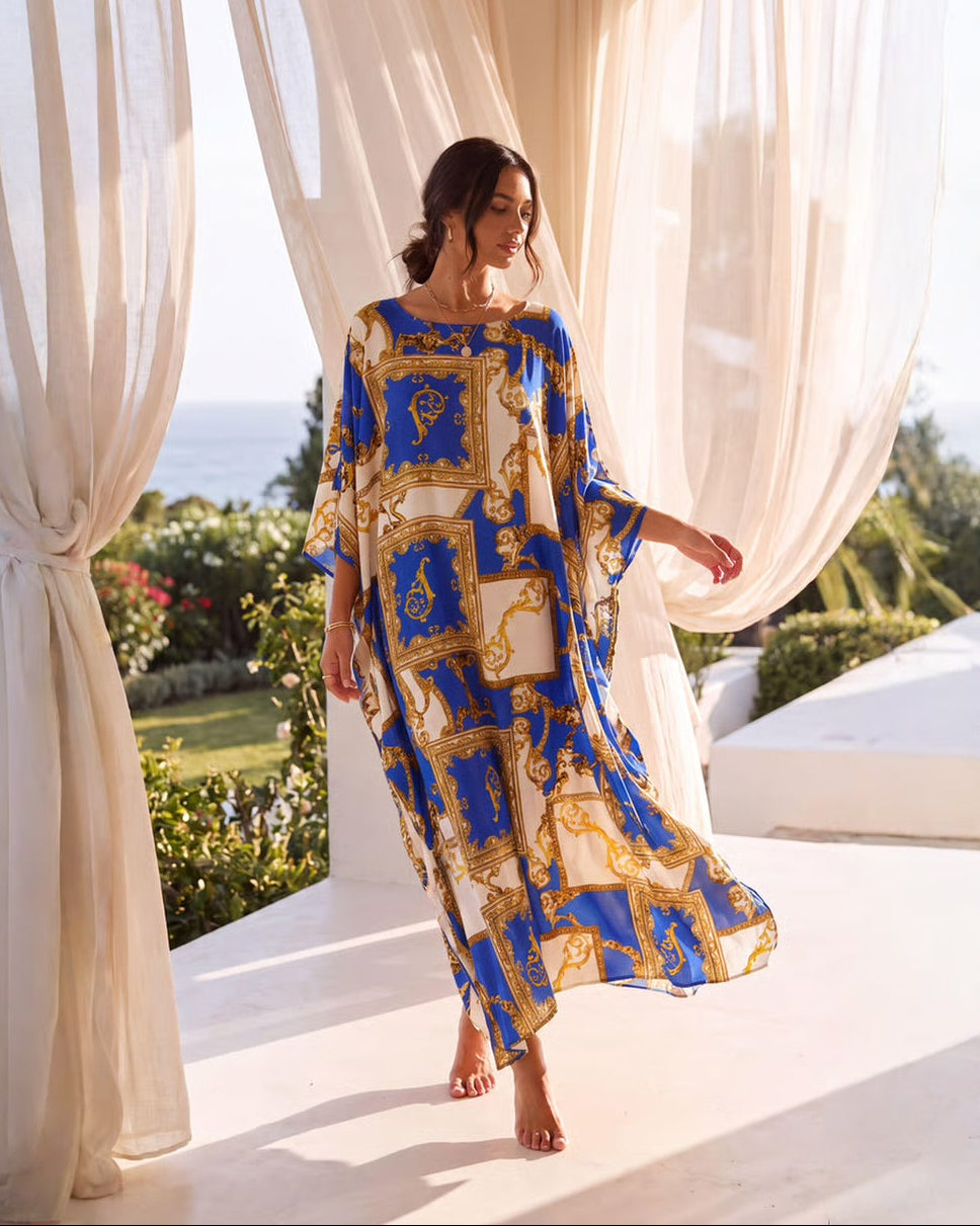 A woman wearing a flowing kaftan dress with a blue, gold, and white baroque-style pattern, standing barefoot on a white terrace framed by sheer cream curtains with a garden and ocean view in the background.