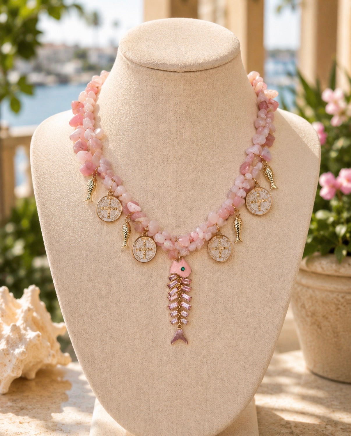 A pink and gold necklace displayed on a beige fabric jewelry bust. The necklace features light pink stone beads, gold-toned fish-shaped charms, round textured medallions, and a dangling fish pendant with pink stones and a small blue eye.