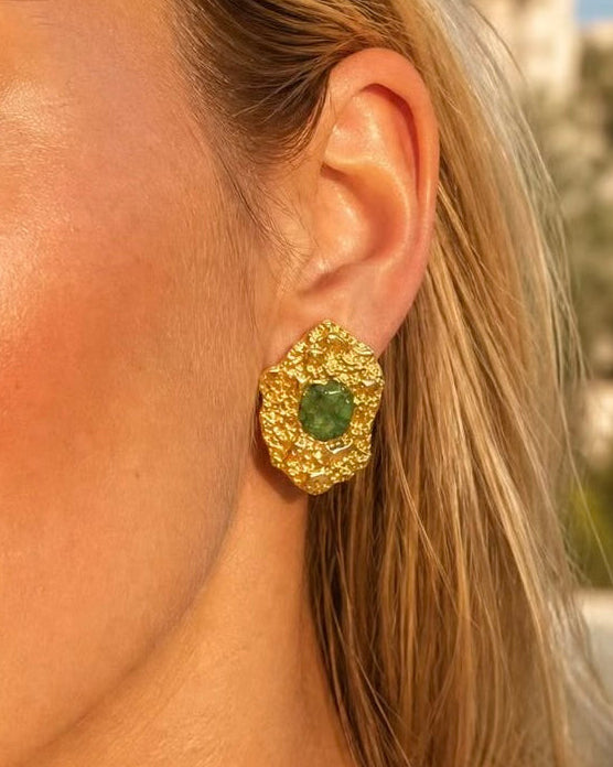 A close-up of a woman wearing a textured gold statement earring with a central green gemstone, set against a softly blurred outdoor background.