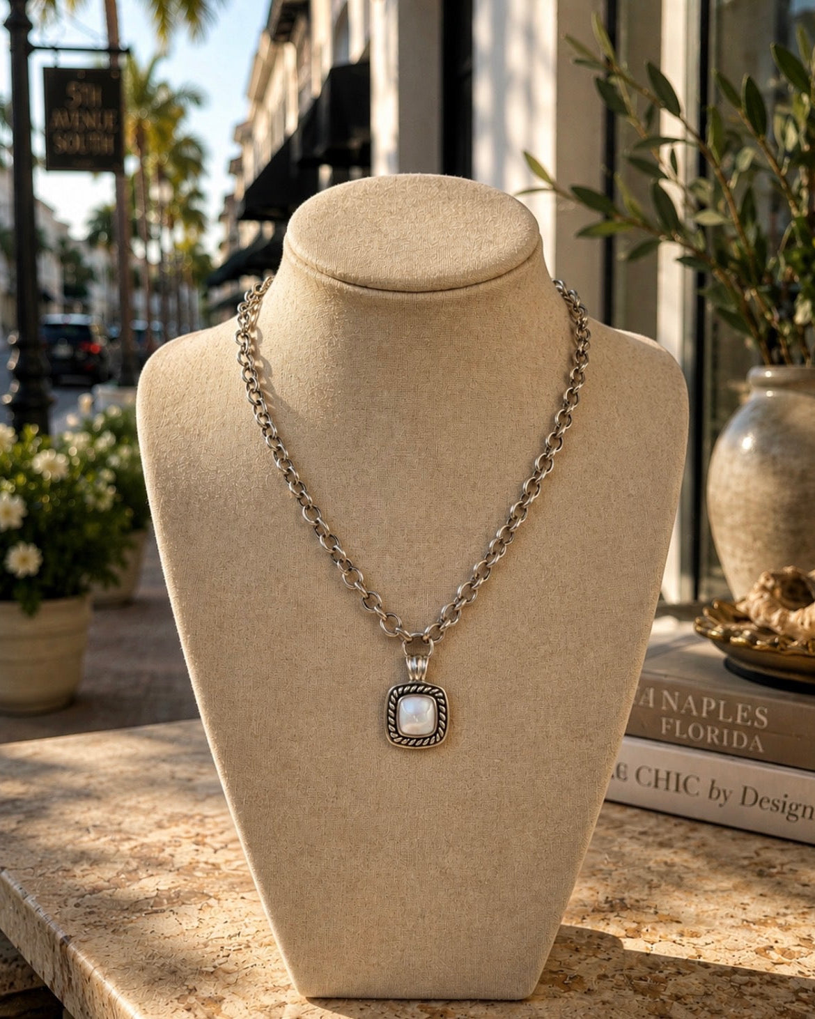 A silver-toned chain necklace with a square pendant featuring a white inlay and dark border, displayed on a beige fabric jewelry bust. The bust is set on a stone counter in a storefront with palm trees and books in the background.