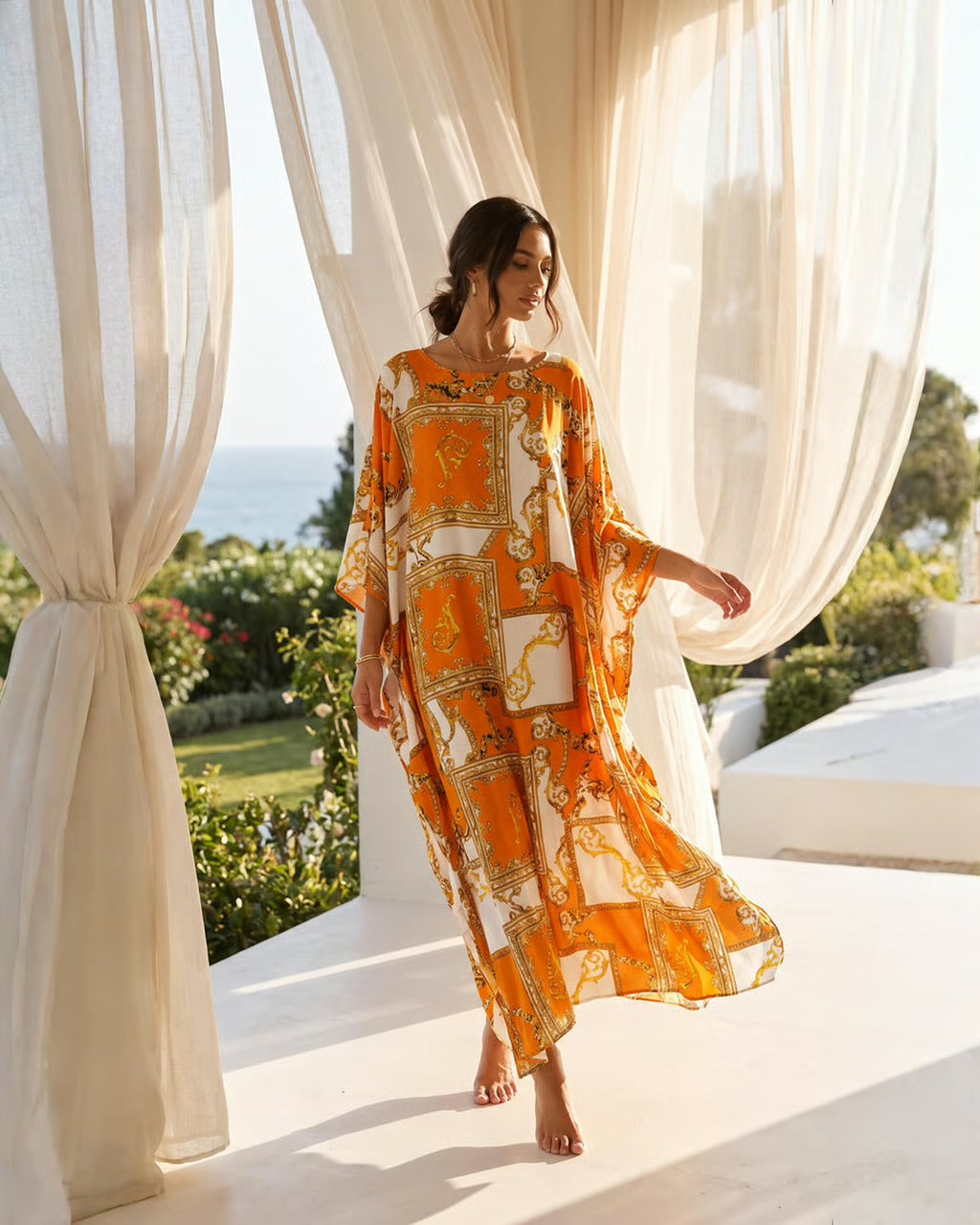A woman wearing a flowing orange and white baroque-patterned kaftan dress walks barefoot on a white terrace framed by sheer cream curtains, with a garden and ocean visible in the background.