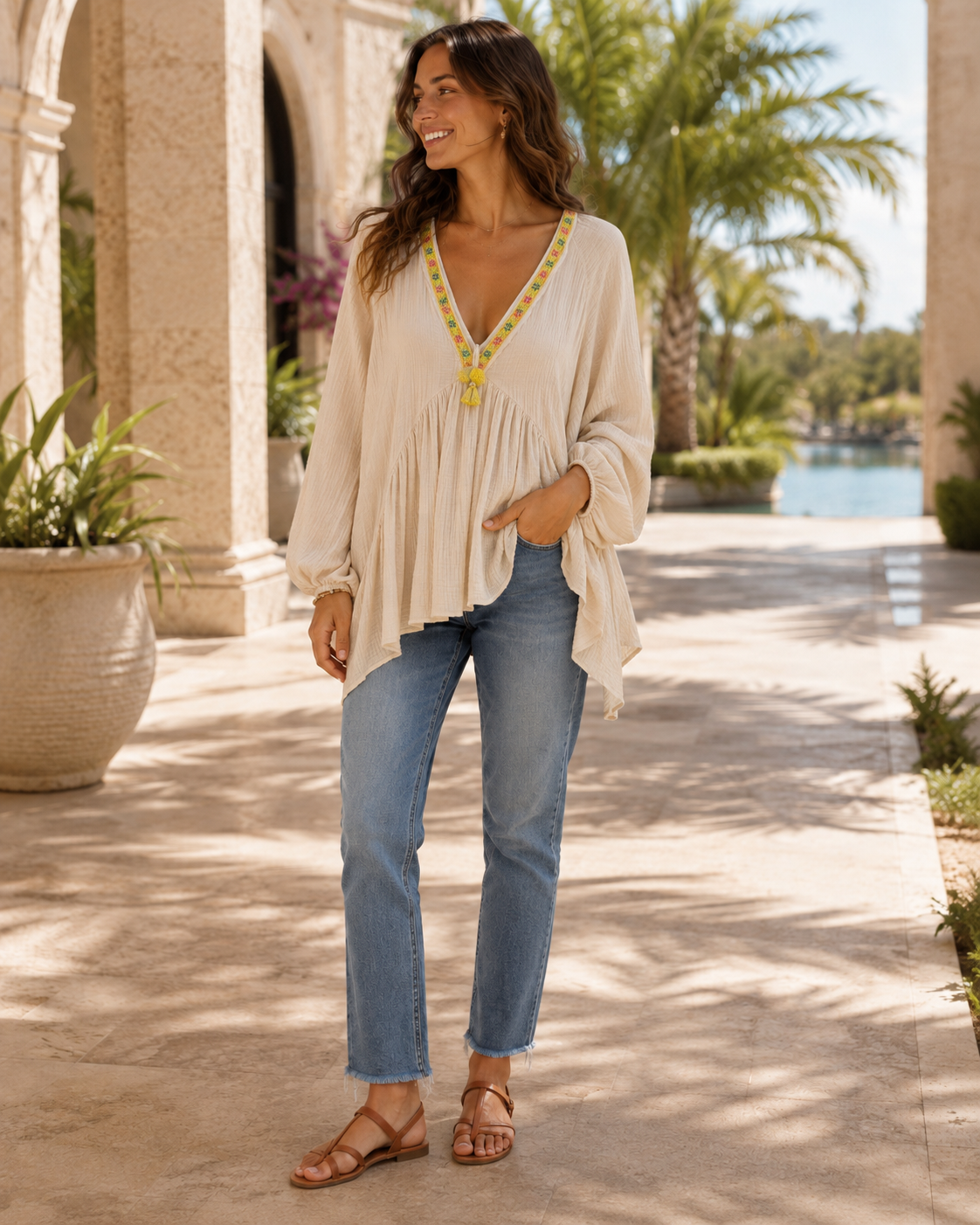 A woman stands outdoors in a sunlit walkway wearing a cream boho-style long-sleeve blouse with yellow and green embroidered trim at the V-neck, paired with light-wash blue jeans and brown strappy sandals.