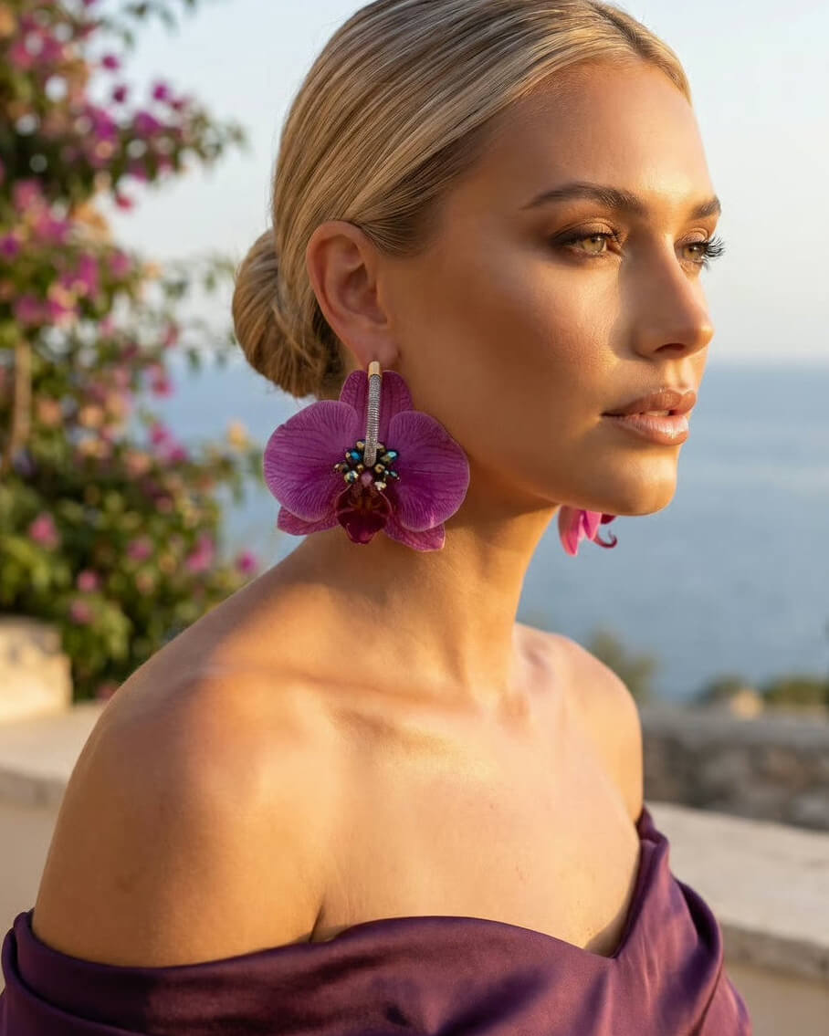 A woman in profile wearing large magenta orchid flower earrings with silver-toned metal details, set against a blurred outdoor background of flowers and water.