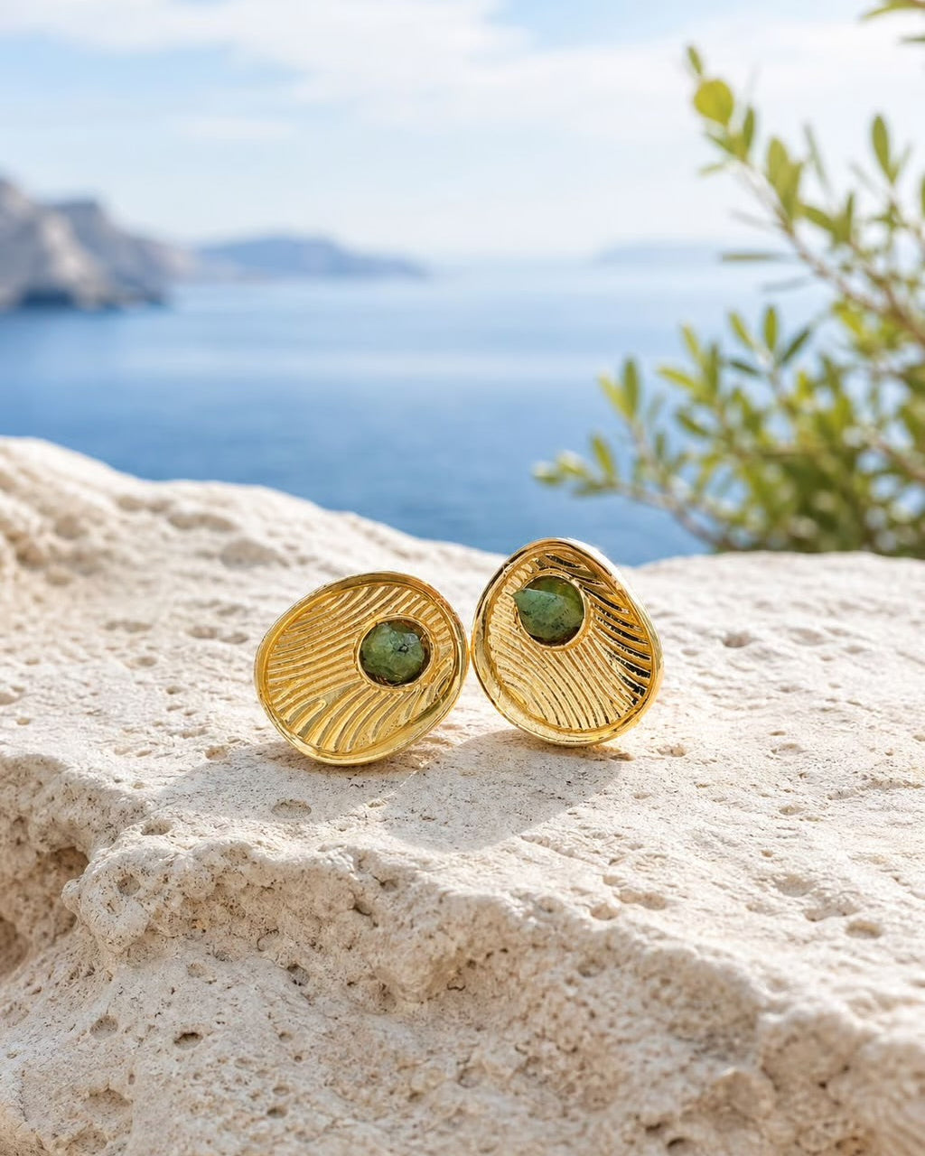 A pair of gold shell-shaped stud earrings with green oval centers, resting on a textured beige stone with a blurred blue sea and sky background.
