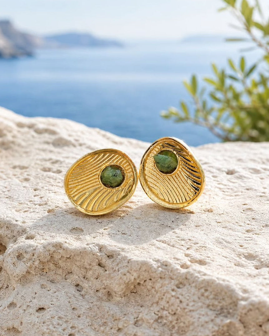 A pair of gold shell-shaped stud earrings with green oval centers, resting on a textured beige stone with a blurred blue sea and sky background.