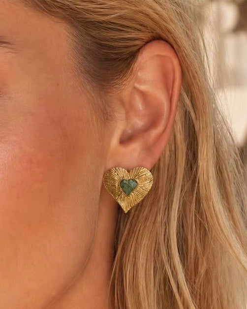 A close-up of a woman's ear wearing a textured gold heart-shaped stud earring with a green heart in the center.