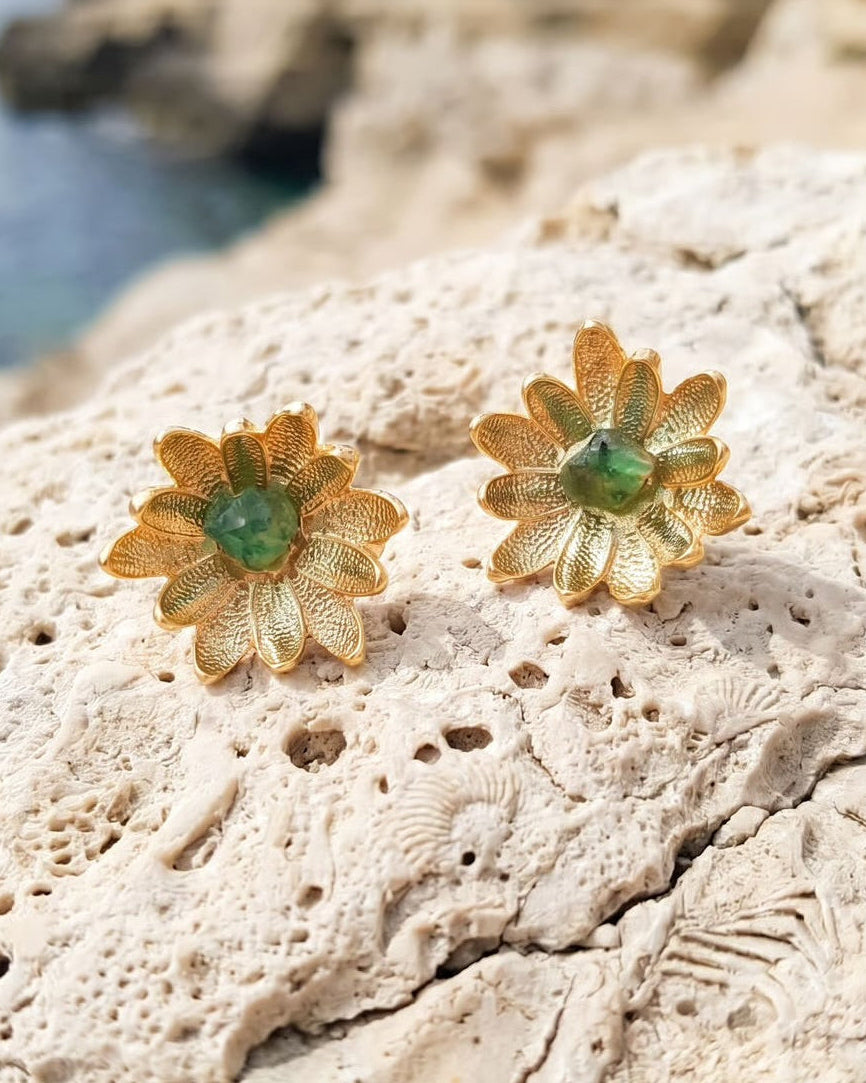 A pair of gold flower-shaped earrings with green gemstone centers, resting on a textured beige rock with a blurred coastal cliff and sea in the background.