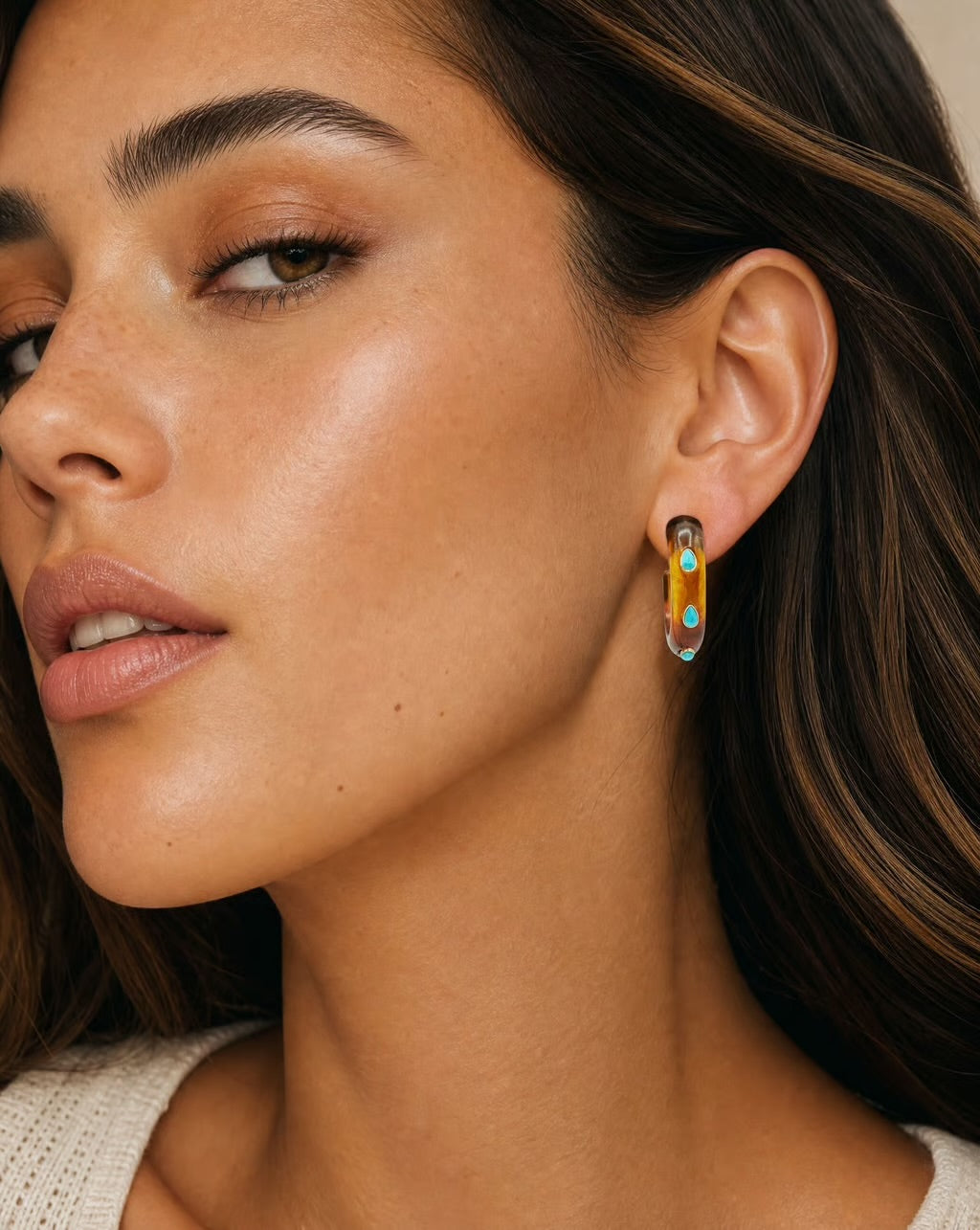 Close-up of a woman wearing a small gold crescent hoop earring with turquoise and blue enamel details.