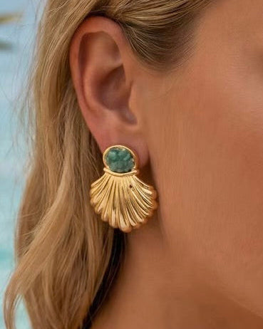 A close-up of a woman's ear wearing a gold shell-shaped drop earring with a round green stone at the top.