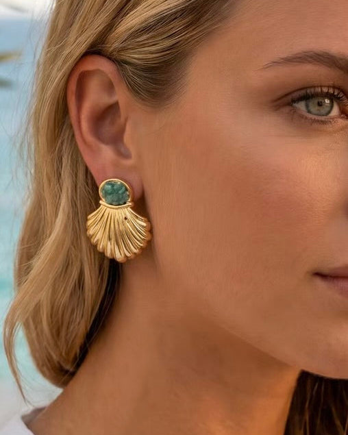 A close-up of a woman's ear wearing a gold shell-shaped drop earring with a round green stone at the top.