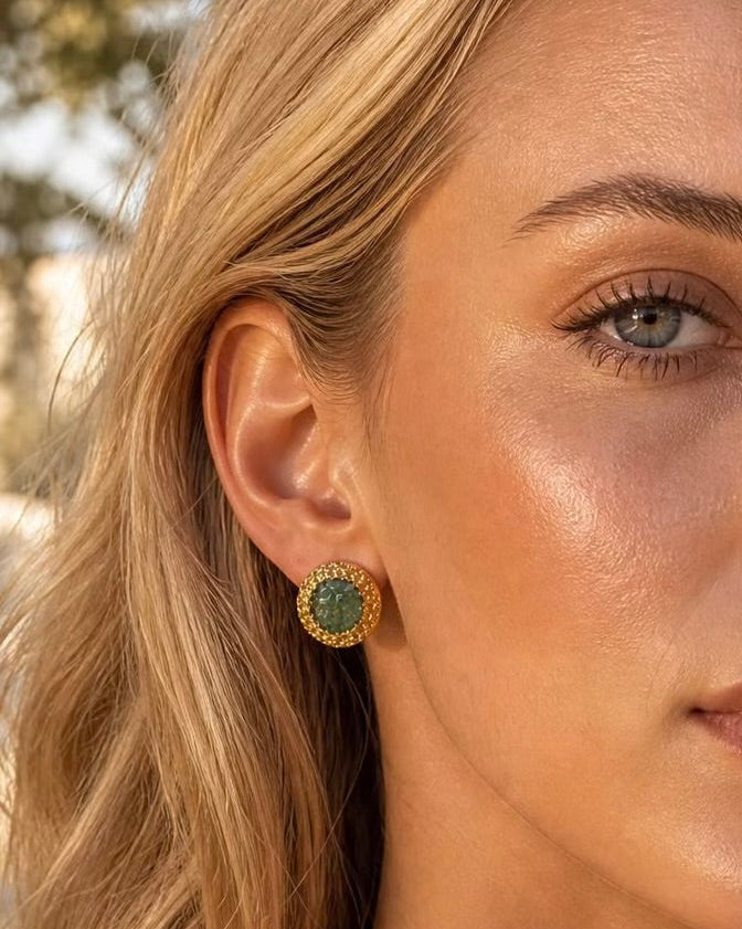 A close-up of a woman's ear wearing a round gold-toned stud earring with a green stone in the center, set against a softly blurred outdoor background.