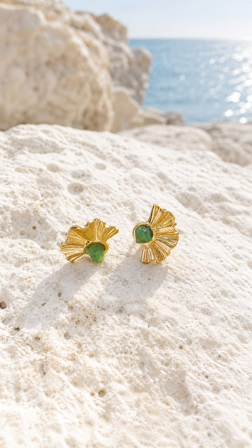 A pair of gold fan-shaped stud earrings with green oval centers, resting on a textured white rock with the blue sea and sky blurred in the background.