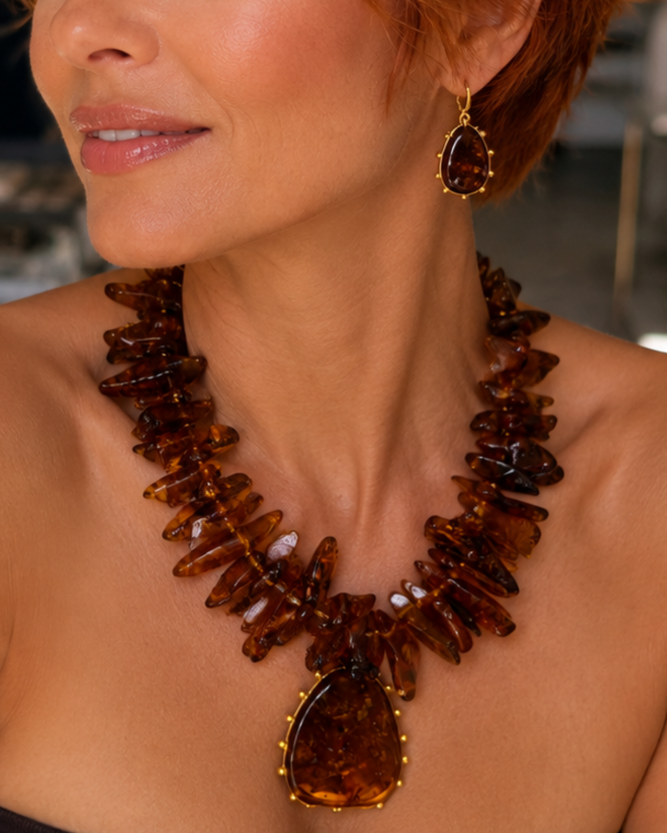 A close-up lifestyle shot of a woman with short red hair wearing a chunky amber-brown resin chunky necklace and matching drop earrings.