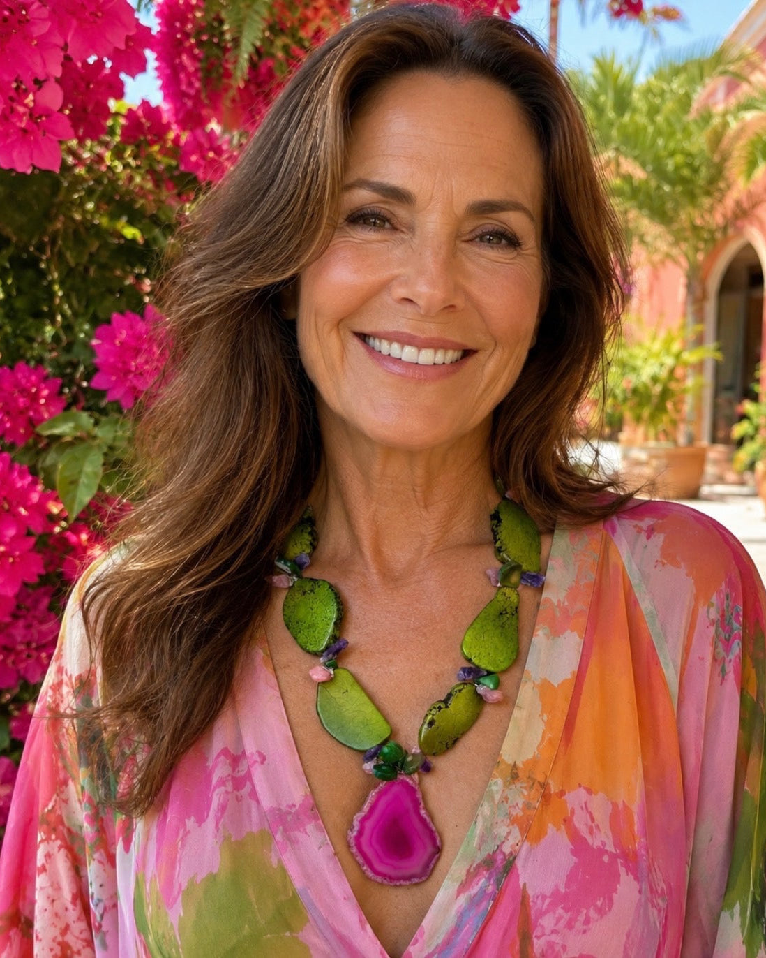 A smiling woman with long brown hair wearing a colorful pink, orange, and green floral top and a chunky green and magenta pendant necklace, standing outdoors in front of bright pink flowers and a warm-toned building.