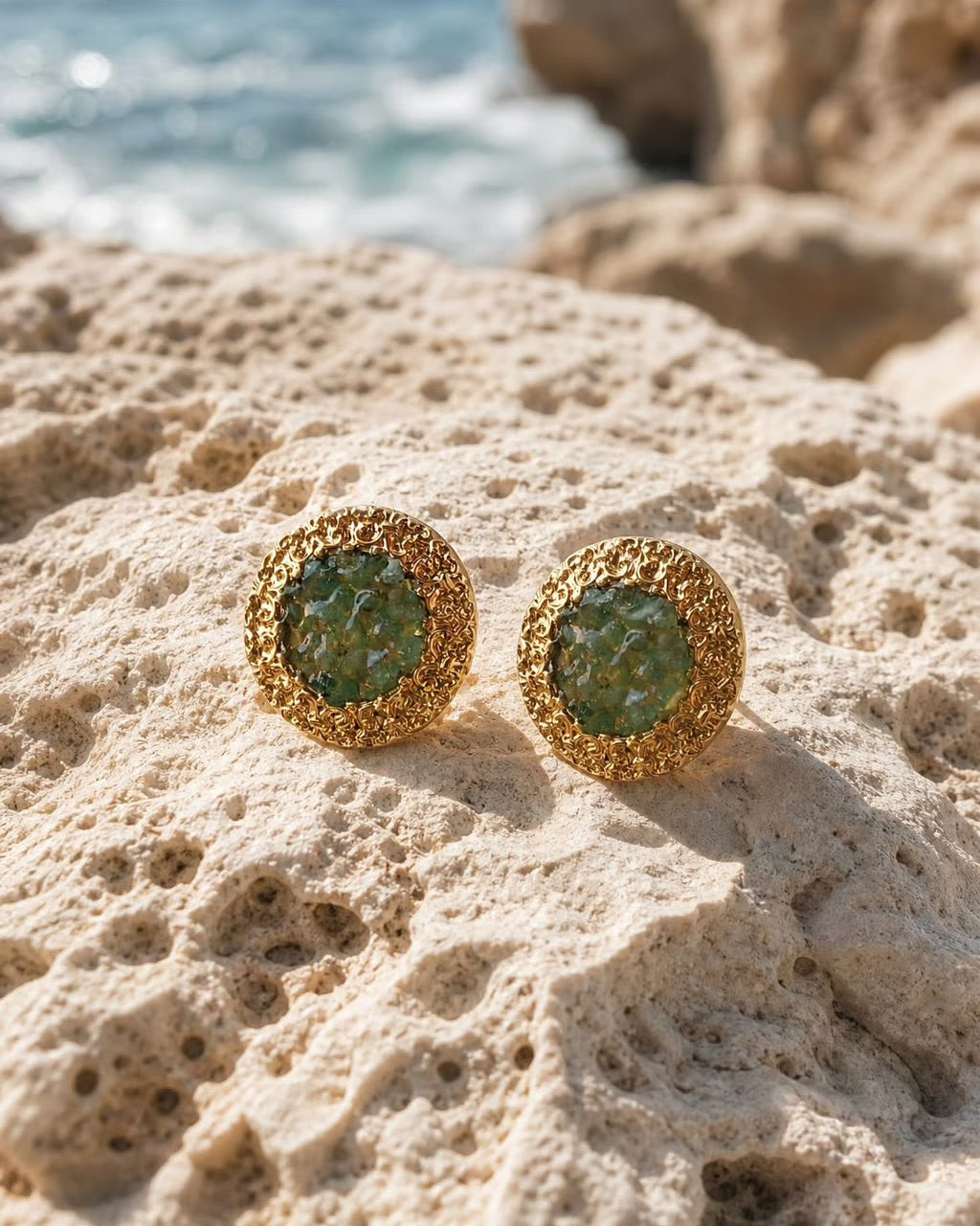 A pair of gold round stud earrings with green stone centers, resting on a textured beige rock with a blurred ocean and cliff background.