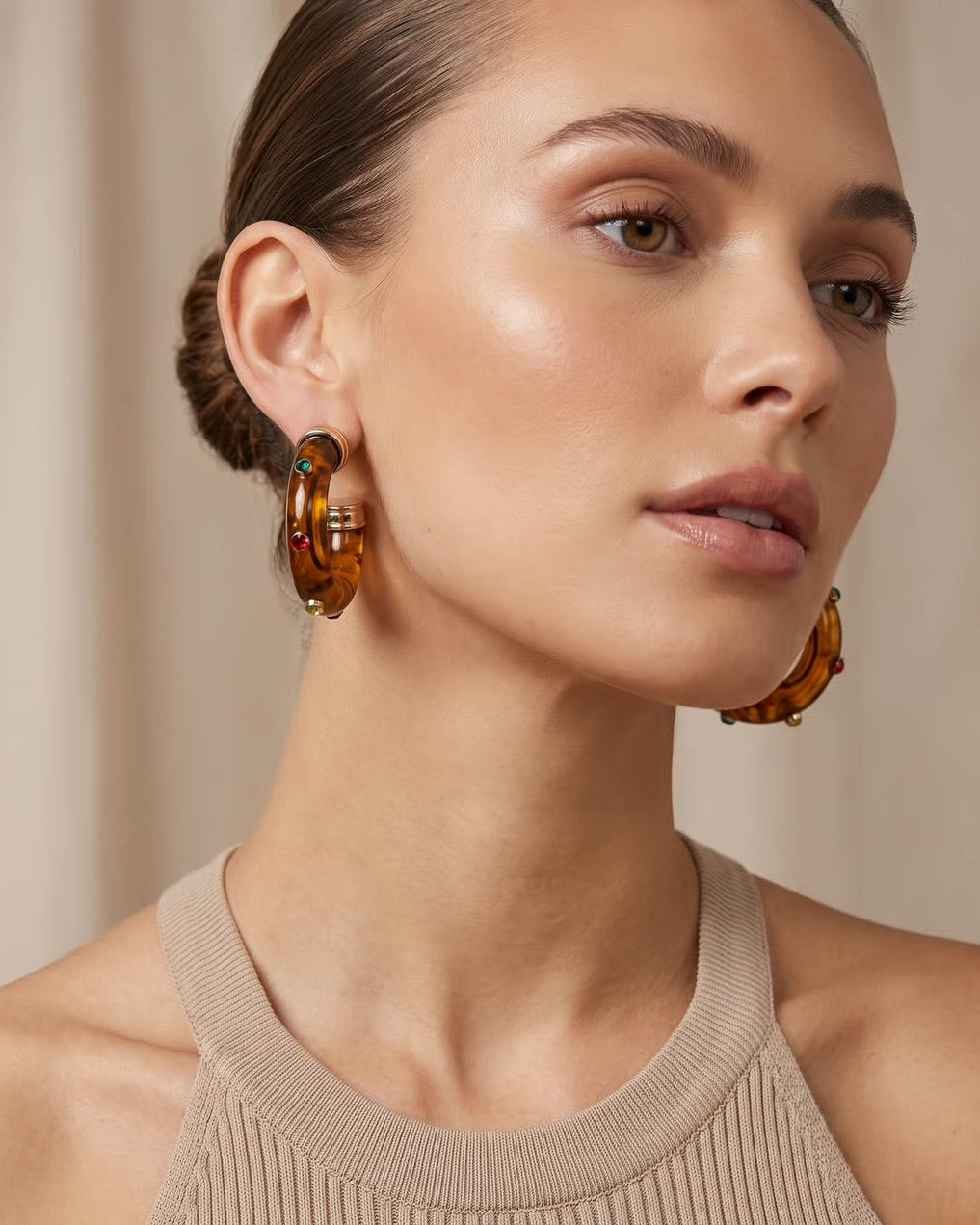 A close-up of a woman wearing large amber-brown hoop earrings with small green and red stone accents, paired with a beige ribbed tank top against a soft beige background.