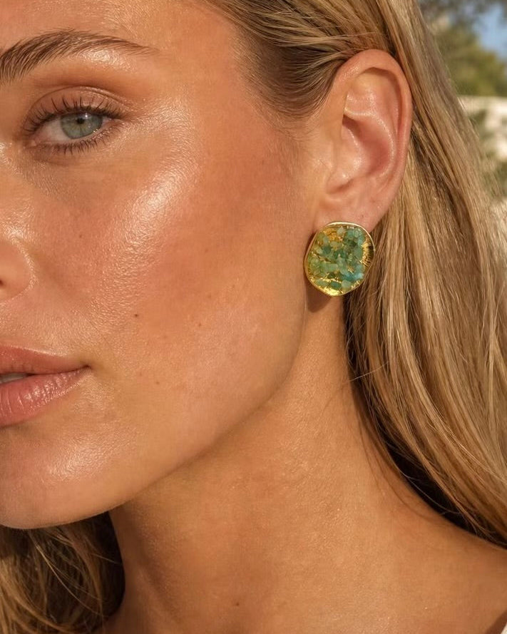 Close-up of a woman wearing a single oval gold-toned earring with a textured green and blue marbled pattern, shown outdoors with natural light.