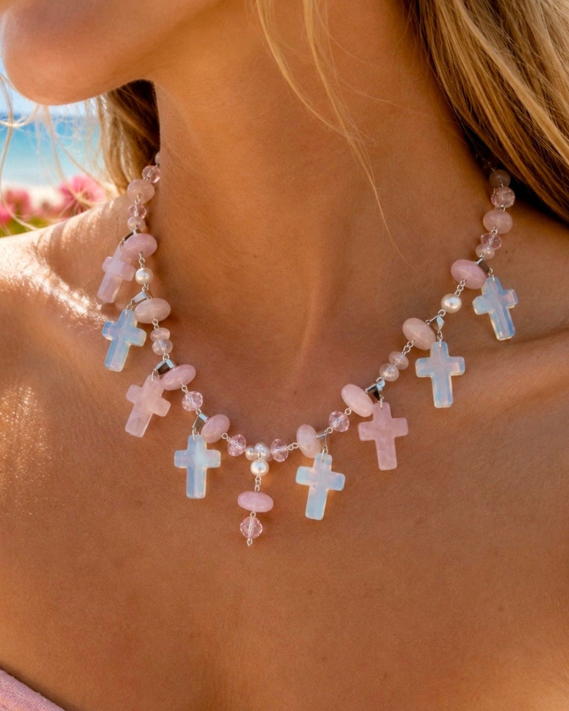 A close-up lifestyle shot of a woman wearing a pink and white beaded necklace with multiple small cross charms, set against a blurred background of pink flowers and the sea.