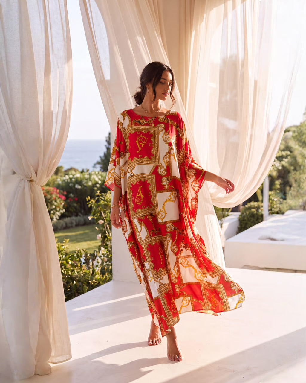 A woman wearing a red, white, and gold baroque-patterned kaftan dress walks barefoot on a sunlit terrace framed by sheer white curtains, with a garden and ocean visible in the background.