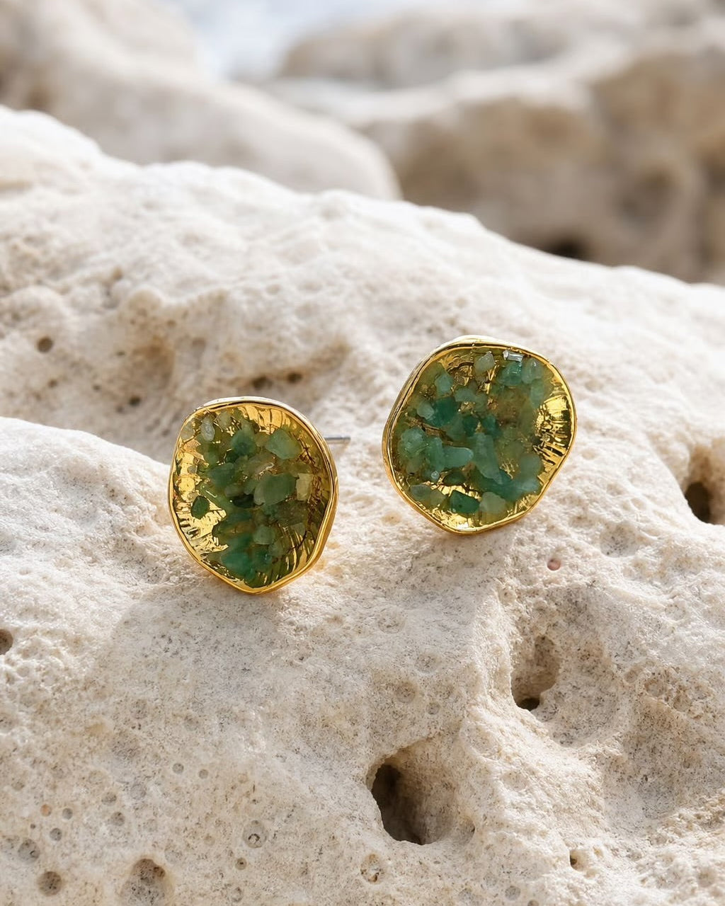 A pair of gold stud earrings with green marbled resin centers, resting on a textured beige rock with a blurred ocean background.