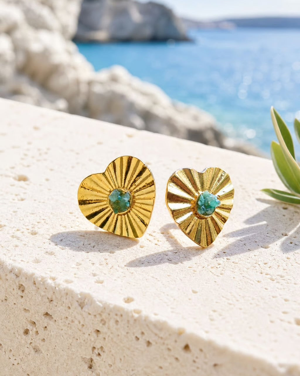 A pair of gold heart-shaped stud earrings with sunburst detailing and a central green stone, resting on a light beige stone ledge with a sunny beach and sea in the background.
