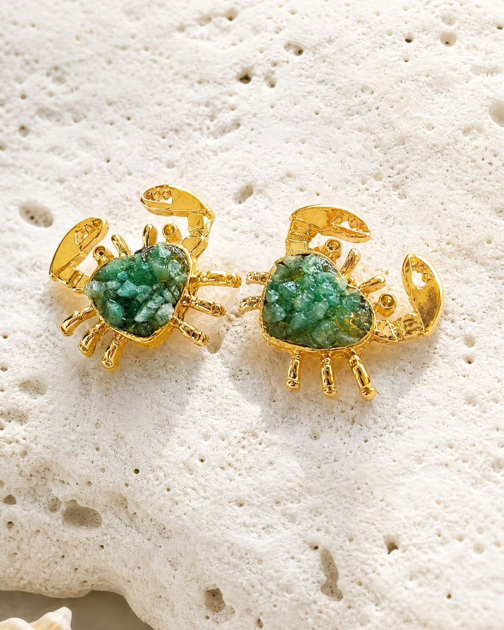 A pair of gold crab-shaped stud earrings with green textured stone centers, displayed on a white textured stone surface with a small seashell nearby.