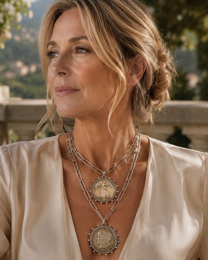 A woman wearing a layered silver necklace with three coin-style pendants, styled with a cream satin top in an outdoor setting.