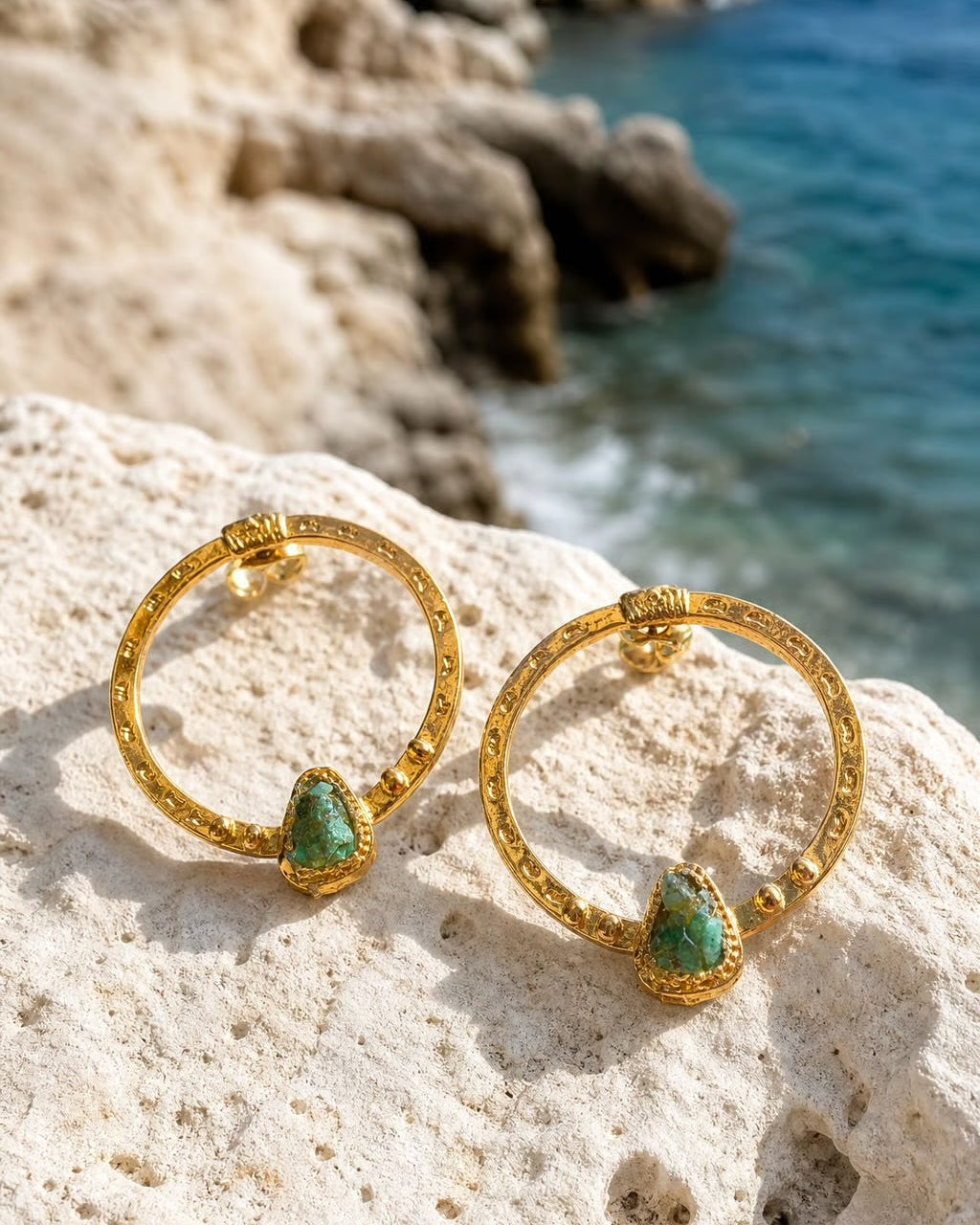 A pair of gold hoop earrings with green teardrop gemstones, resting on a white rock with a coastal cliff and blue sea in the background.