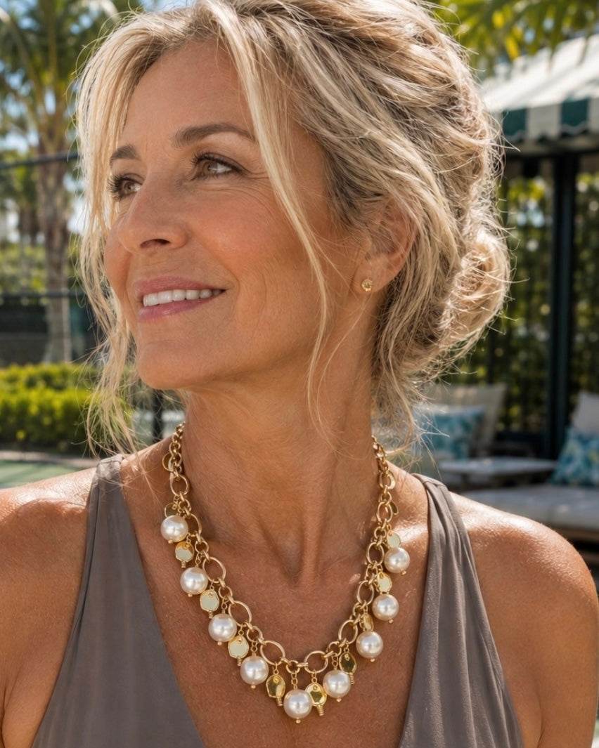 A woman wearing a gold chain necklace with white faux pearls and gold spacer rings, paired with a taupe halter top, photographed outdoors near a poolside lounge area.