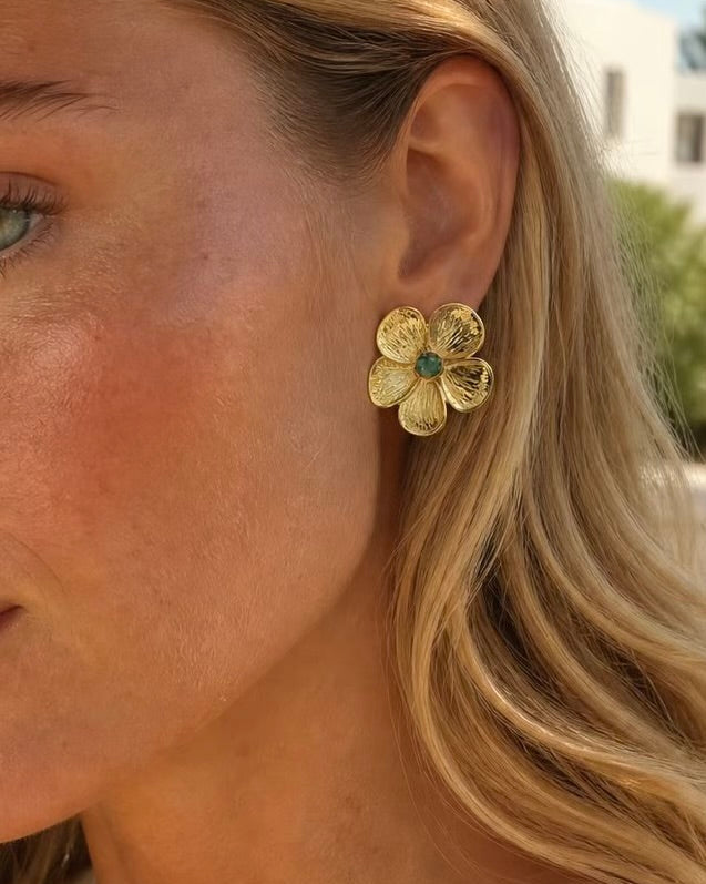 A close-up of a woman's ear wearing a textured gold flower-shaped stud earring with a small turquoise blue center, set against an outdoor background.