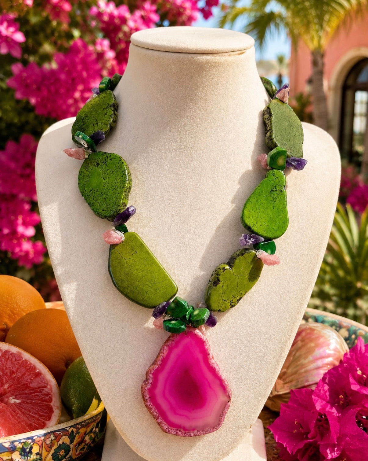 A colorful statement necklace made of green stone slices and a bright pink agate pendant, displayed on a cream-colored mannequin bust with a tropical garden background.